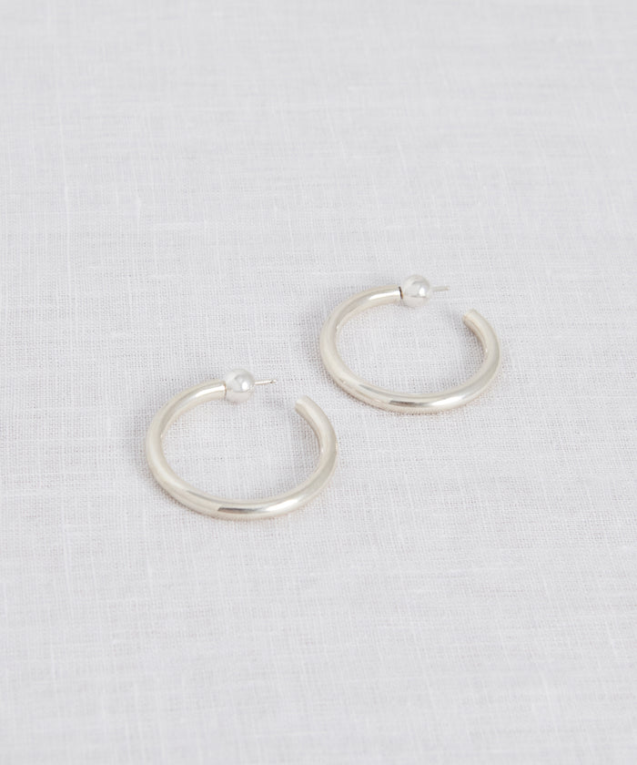 The Small Clara Hoops, crafted in sterling silver with post backings, are displayed on a light, textured fabric surface.