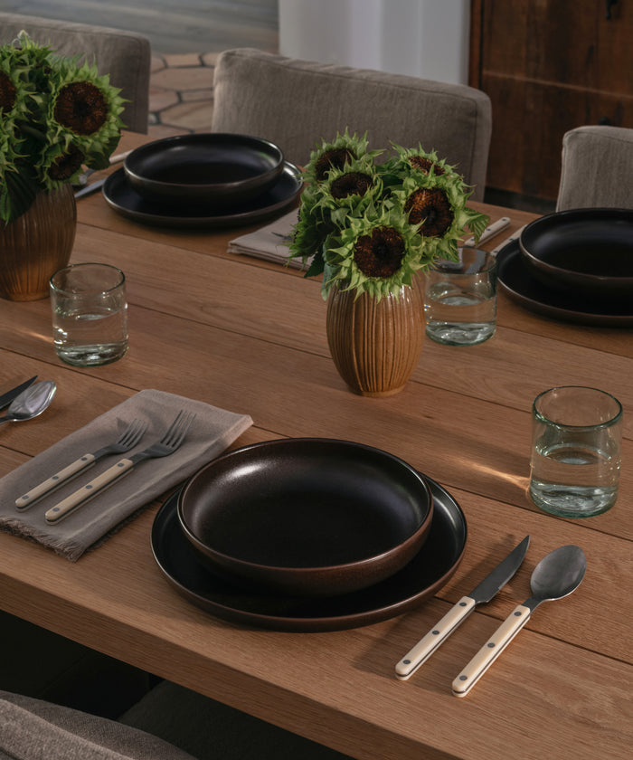 A dining table set with ceramic dinnerware and recycled glass cups with vases filled with florals.