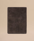 The Suede Passport Holder, a travel essential in dark brown with a soft, textured finish and subtle embossed text near the bottom, is centered on a light beige background.