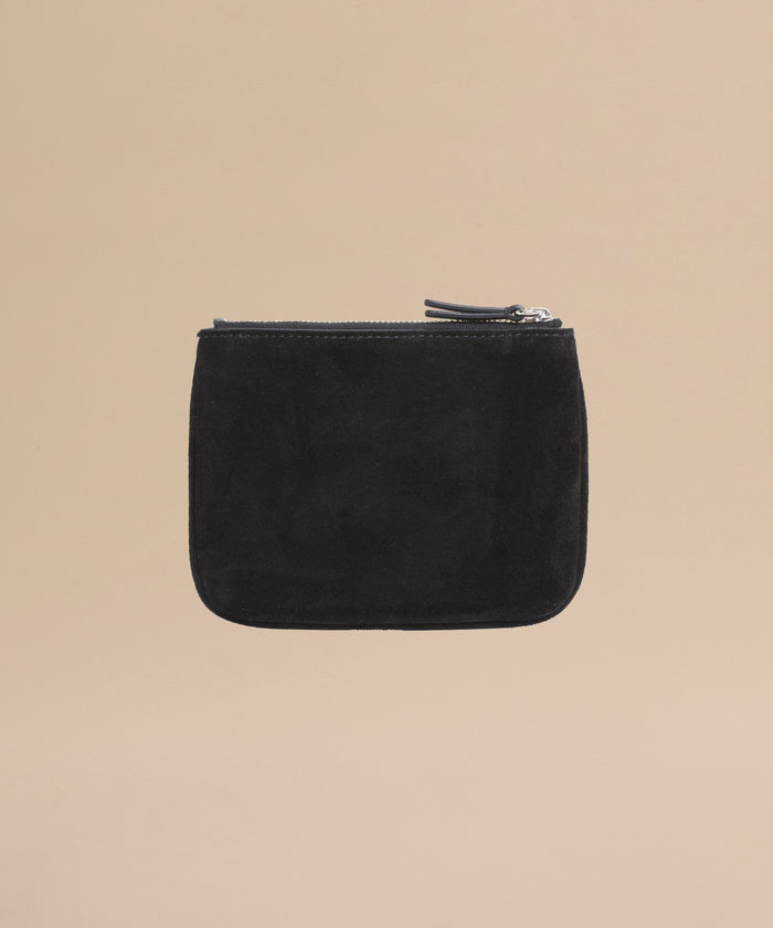 The Suede Pouch is a small black suede accessory with a zippered closure and minimalist design, perfect for use as a compact pouch or passport holder, pictured on a plain beige background.