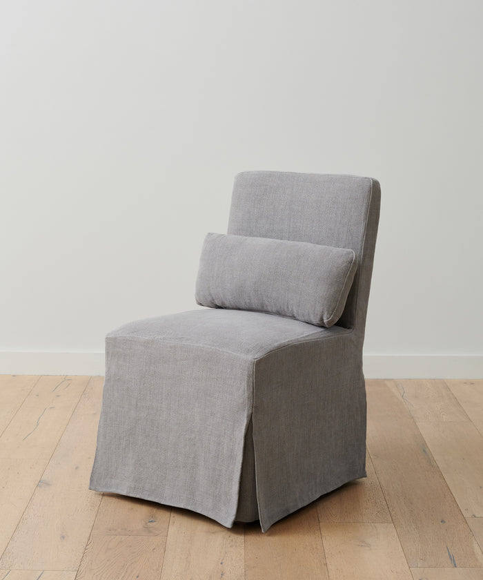 The Sunset Dining Chair Slipcover in ash linen covers a modern armless chair, paired with a matching rectangular cushion and set on a light wood floor against a plain white wall.