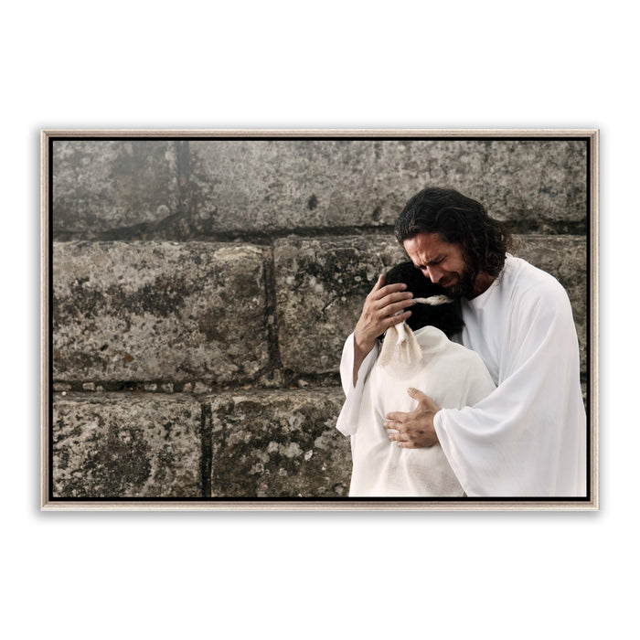 Color photograph of Jesus on the right in a white robe, embracing a woman in white whose face is buried against His shoulder. His face is visible, brow furrowed, weeping with her. Stone wall background, daylight.