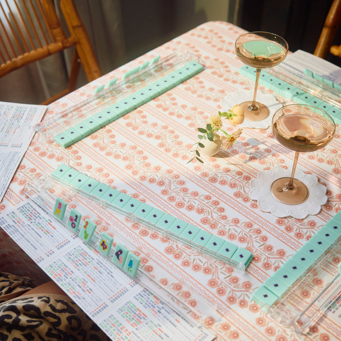 A tabletop game set with mint green domino tiles arranged on a patterned tablecloth, alongside two filled coupe glasses, a small floral centerpiece, and printed game cards.