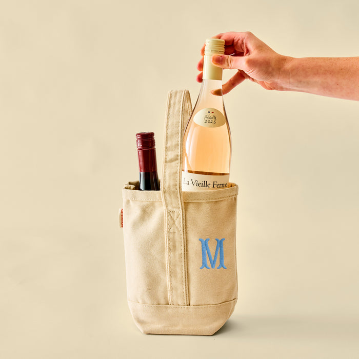 A hand placing a bottle of rosé wine into a cream canvas tote bag that features a blue embroidered 'M' monogram.