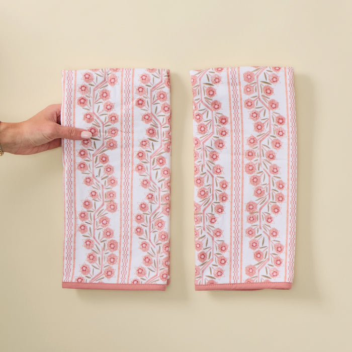A top-down view of two folded kitchen towels resting side-by-side on a cream background. The towels feature a pattern of vertical stripes and pink and peach floral climbing vines, finished with a solid pink border at the bottom. A person's hand is visible touching the left edge of the towel on the left.