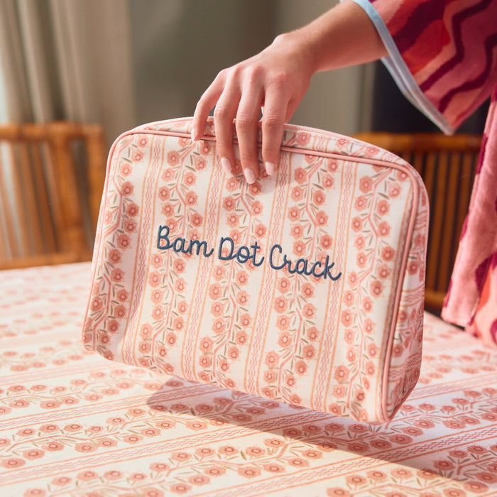A hand holding a rectangular toiletry bag featuring a pink and coral floral stripe pattern with the phrase 'Bam Dot Crack' embroidered in dark blue, set against a matching patterned fabric background.