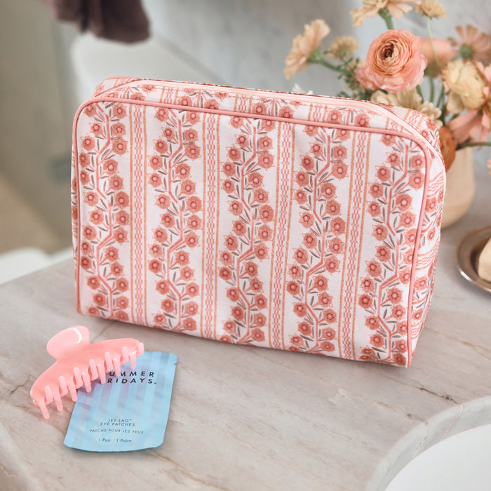 A large toiletry bag featuring a pink and coral floral climbing vine pattern with pink piping, sitting on a light marble surface. In the foreground is a pink hair claw clip resting on a blue Summer Fridays eye patch packet, with a vase of pink flowers blurred in the background.