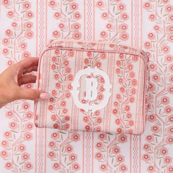A hand holds a pink and white floral patterned toiletry bag featuring a large white applique monogram 'B' on the front, set against a matching patterned background.