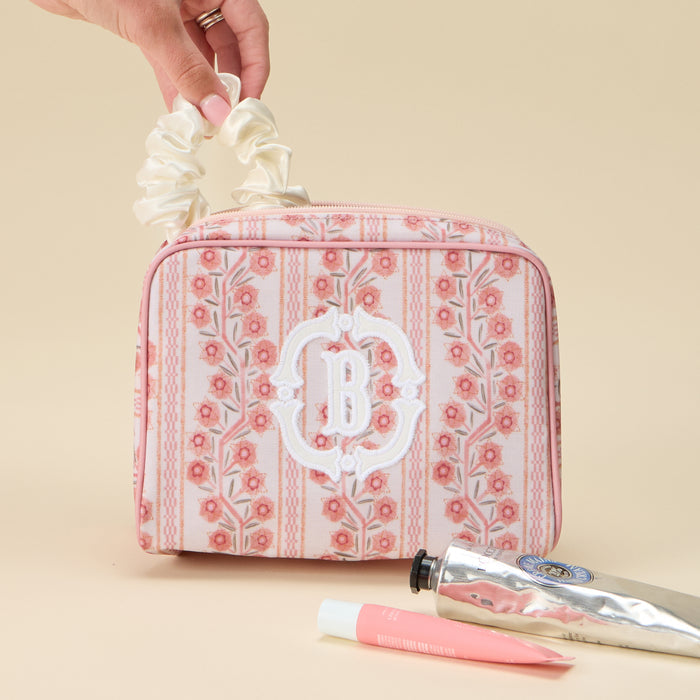 A hand holding a white scrunchie above a pink and coral floral striped toiletry bag featuring a large white applique monogram 'B' on the front. The bag is set on a light beige surface next to two tubes of lotion or cream.