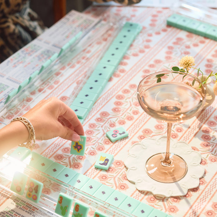 A hand plays with mint green mahjong tiles arranged on a patterned tablecloth next to a coupe glass and a small flower sprig.