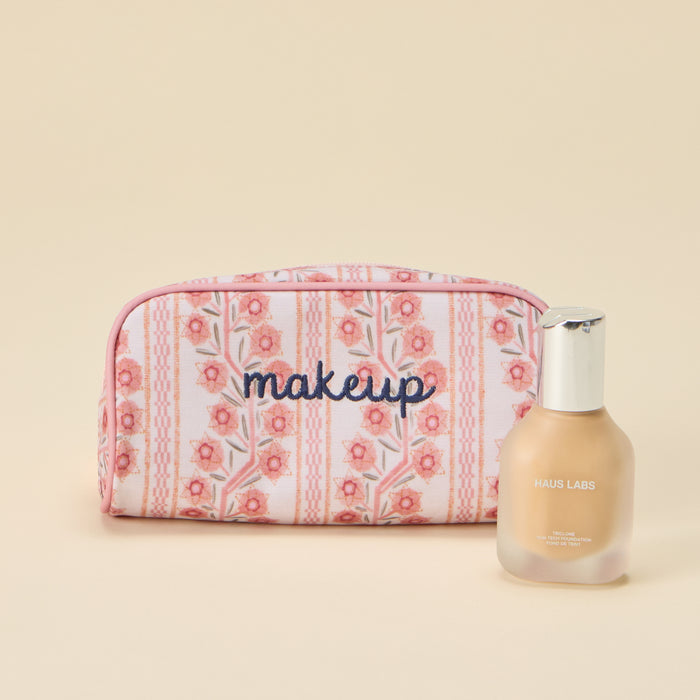 A rectangular toiletry bag featuring a pink and coral floral climbing vine stripe pattern with light pink piping, with the word 'makeup' embroidered in dark blue cursive across the front. A glass bottle of Haus Labs liquid foundation sits next to the bag against a plain light background.