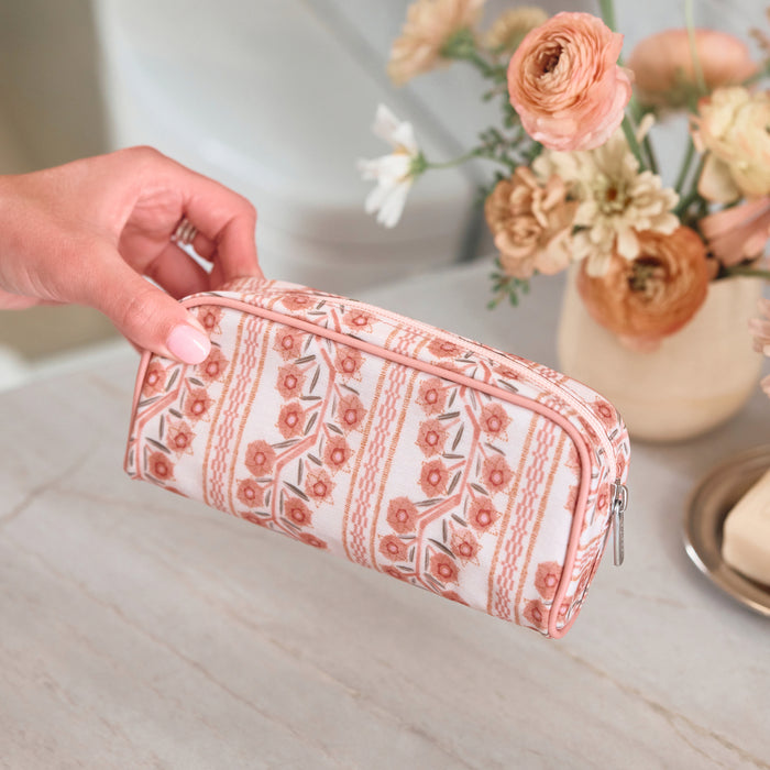 A hand holding a small toiletry bag with a pink and cream floral climbing vine pattern, placed on a light marble surface next to a vase of pink flowers.