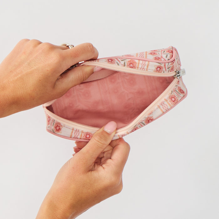 A close-up of hands holding open a small zippered toiletry bag to reveal a solid pink interior lining. The exterior of the bag features a delicate pink and green floral climbing vine pattern against a plain light background.