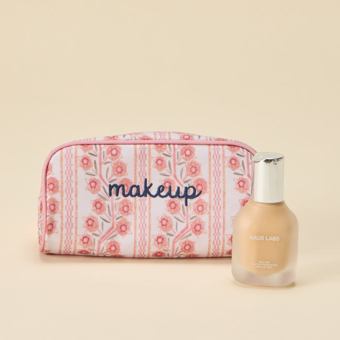 A small toiletry bag with a pink floral pattern and the word 'makeup' embroidered in dark blue cursive on the front. The bag is positioned next to a bottle of Haus Labs foundation against a solid light beige background.