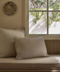Two Adler Lumbar Pillows in beige linen blend sit on a plaid-cushioned bench by the window, sunlight casting leafy shadows nearby. Above the pillows, a round textured wall decor adds interest to the serene space.