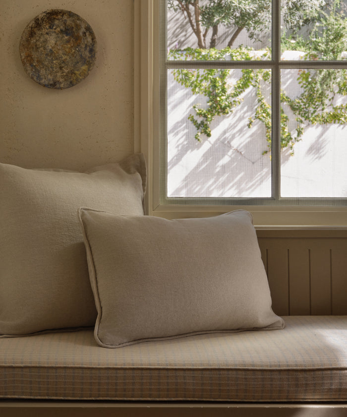 Two Adler Lumbar Pillows in beige linen blend sit on a plaid-cushioned bench by the window, sunlight casting leafy shadows nearby. Above the pillows, a round textured wall decor adds interest to the serene space.