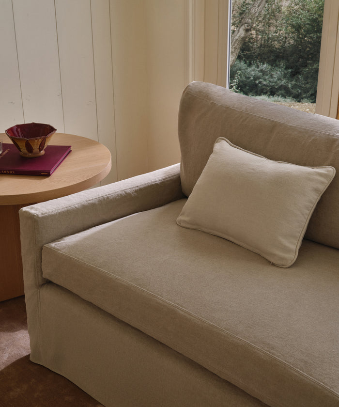A beige armchair with a matching Adler Lumbar Pillow sits beside a round wooden table that holds a red bowl and a closed burgundy book near a window overlooking greenery.