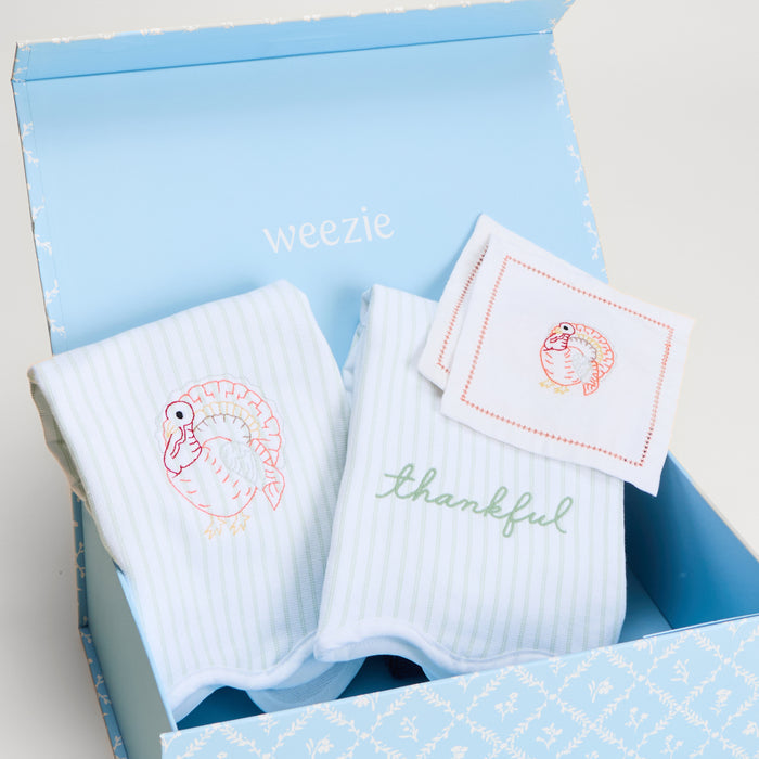 A gift set in a light blue box from Weezie, featuring two green striped kitchen towel and square cocktail napkins with embroidered turkey designs, all arranged in the box.
