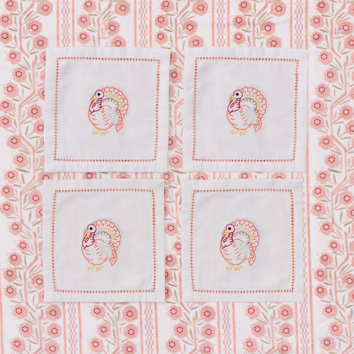 Four square white napkins with embroidered turkeys arranged in a 2x2 grid on a pink and white patterned fabric background.