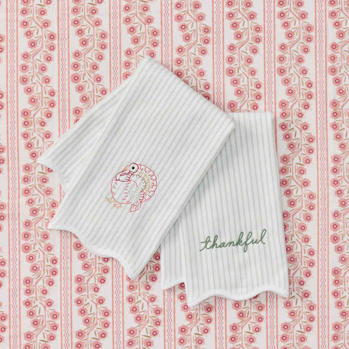 Two green striped kitchen towels with scalloped edges, featuring a pink and green embroidered turkey on one towel and the word 'thankful' in green script on the other, displayed on a pink and white patterned background.