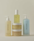 Five Oak Essentials’ The Body Routine, featuring a spray bottle, two pump bottles, and two jars with minimalist labels, is arranged neatly on a light beige background in soft yellow and blue tones.