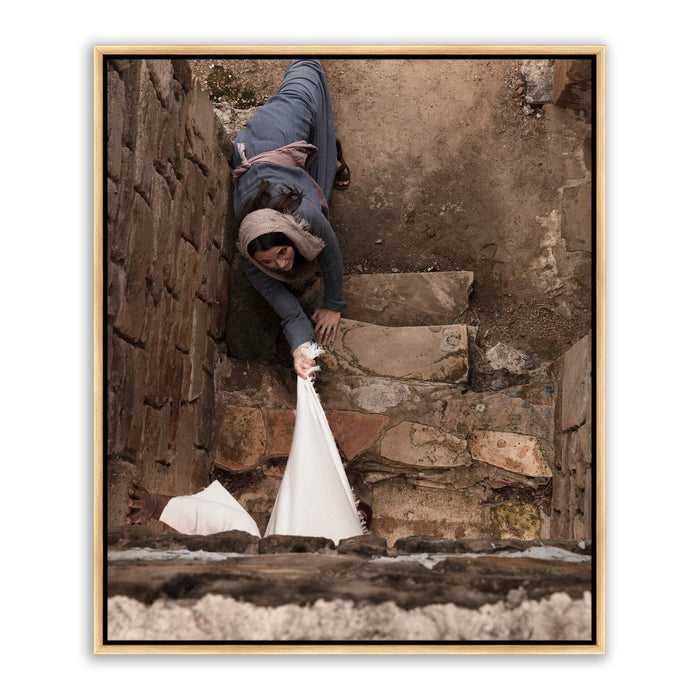 Aerial high-angle photograph looking down a stone staircase. A woman in a gray robe lies on the steps, arm extended, fingers reaching toward the visible hem of a white robe at the bottom edge of the frame.