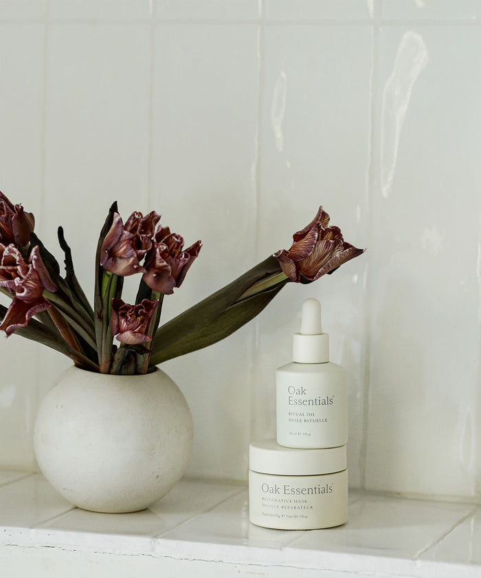A white vase with dried purple flowers sits next to The Reset Duo—two Oak Essentials skincare essentials—against a white tiled background, perfect for elevating your skincare routine.