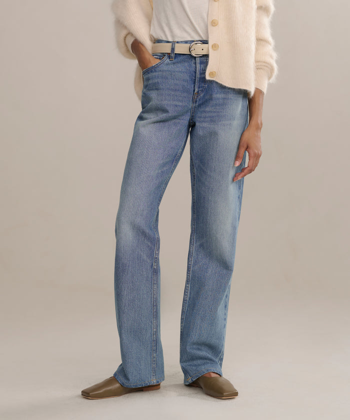 A person stands against a plain light background wearing The Standard higher mid-rise straight leg jeans, a white belt, white t-shirt, cream cardigan, and brown flat shoes.