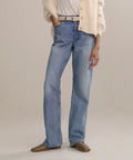 A person stands against a plain light background wearing The Standard higher mid-rise straight leg jeans, a white belt, white t-shirt, cream cardigan, and brown flat shoes.