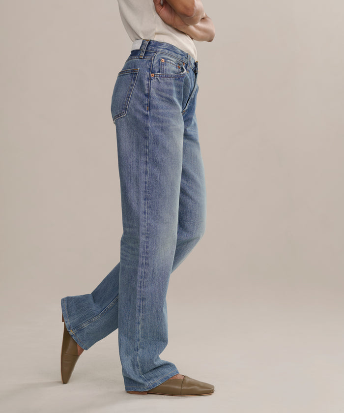 Wearing The Standard higher mid-rise blue jeans, a person in a white shirt and brown flat shoes stands with arms crossed against a plain light background; only the lower half is visible.