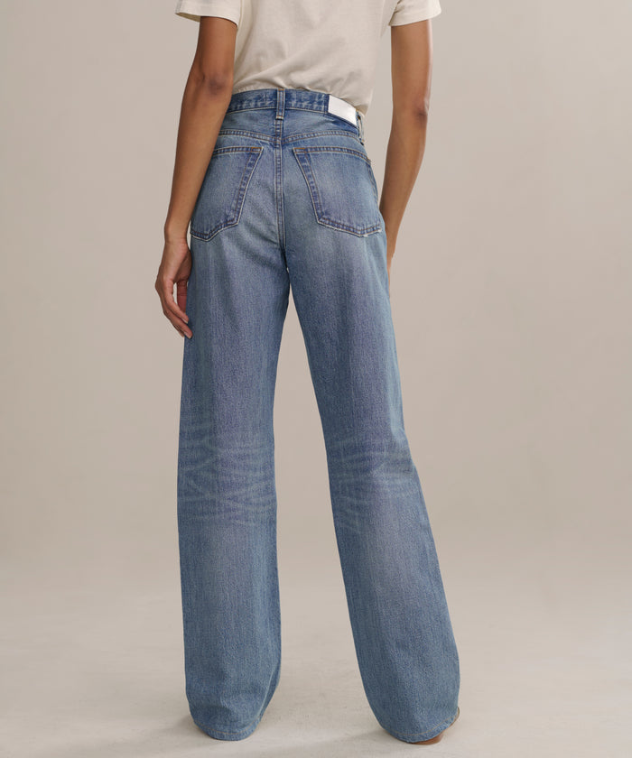A person stands on a light surface facing away, wearing The Standard: cream short-sleeve shirt and higher mid-rise, straight-leg jeans with visible back pockets and seams.
