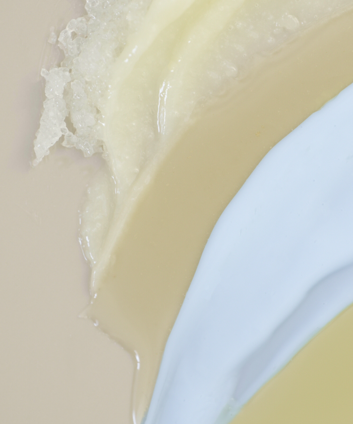 A close-up of The Travel Body Routine’s three creamy formulas—off-white, beige, and light blue—smeared in textured horizontal layers, ideal for your travel skincare needs.