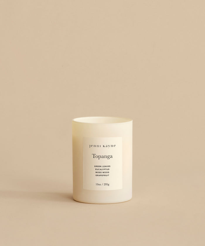 A Topanga Glass Candle rests in a minimalist white container on a beige background. The label reads jenni kayne, Green Leaves, Blackwood, Grapefruit. This natural candle is hand-poured in Los Angeles, 10oz / 295g.