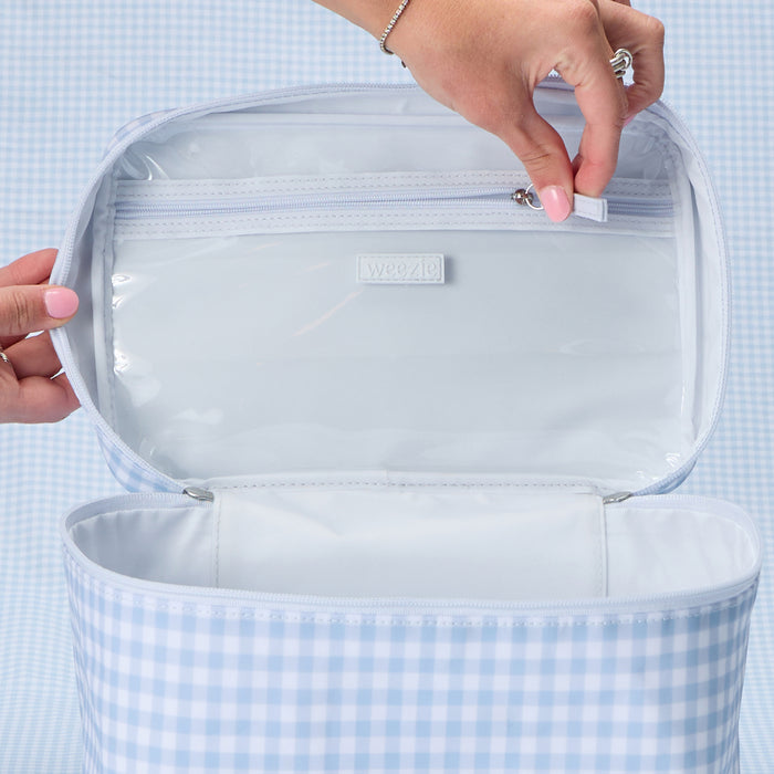 Close-up view of the interior of a light blue and white gingham vanity case, showing a hand opening the zippered pocket on the underside of the lid, which features a 'Weezie' brand label.
