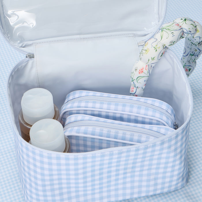 Open light blue and white gingham vanity case containing two matching smaller toiletry bags and two bottles of skincare products, with a floral-patterned headband resting on the rim of the case.