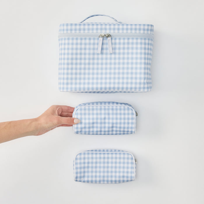 Top-down view of the Weezie Travel Bundle in light blue and white gingham, featuring one large vanity case at the top and two small matching toiletry bags below, with a hand positioned to touch the middle bag.