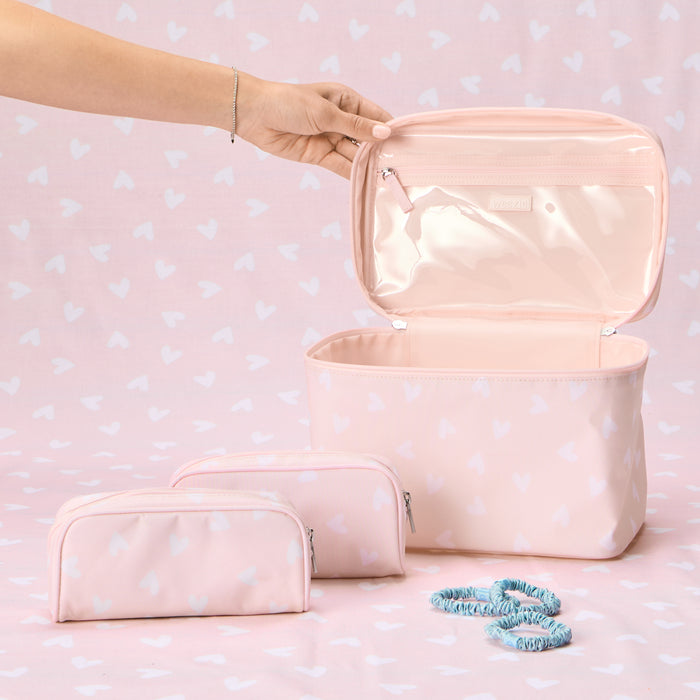 A Weezie Travel Bundle in a Tossed Pink Hearts pattern, featuring one large, open vanity case with a clear interior lid pocket and two matching small toiletry bags, arranged on a pink heart-patterned background with three blue scrunchies.