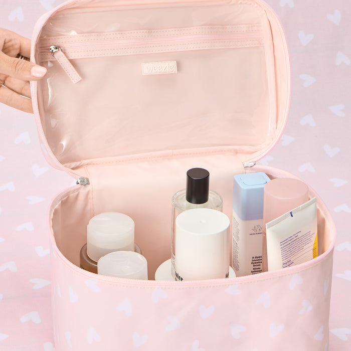 Close-up of the interior of an open vanity case in a 'Tossed Pink Hearts' pattern, showing a hand pulling open the clear, zippered pocket on the underside of the lid, with various skincare bottles standing inside the main compartment.