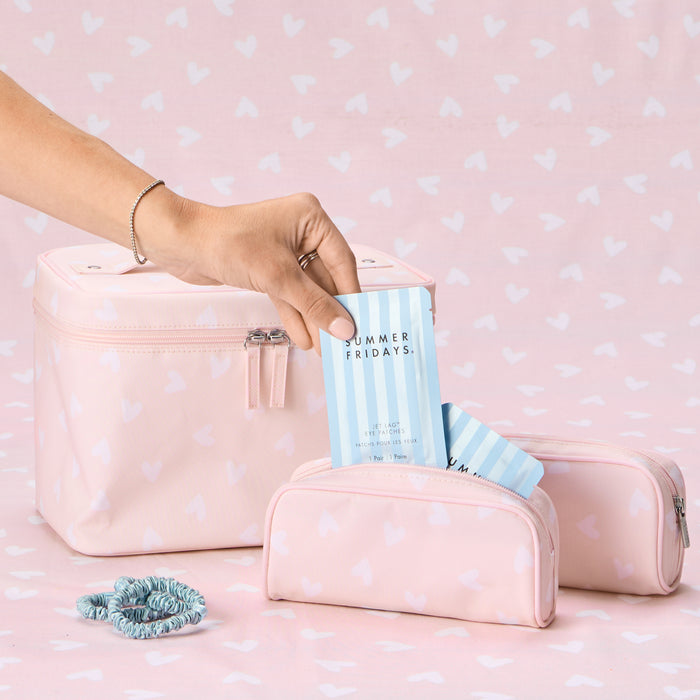 A hand placing a Summer Fridays eye patch packet into a small Tossed Pink Hearts toiletry bag, with a larger matching vanity case and a second small bag nearby on a heart-patterned background, accompanied by blue hair scrunchies.