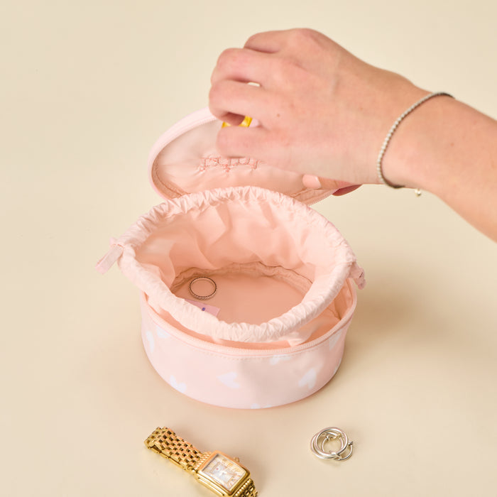 A round blush pink jewelry organizer with a drawstring-lined interior, shown open with a hand lifting the lid. A gold watch and silver rings are placed nearby on a warm beige surface.