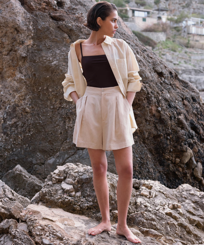 A woman stands barefoot on rocky terrain, layering a light button-up over the Tre One Piece swimsuit and beige high-waisted shorts, hands in her pockets as she gazes thoughtfully to the side.