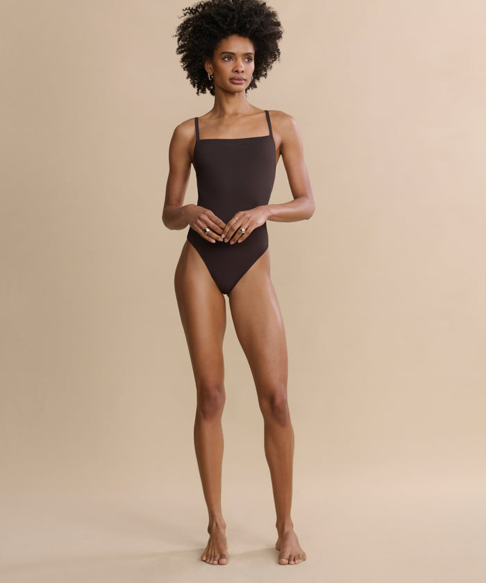 A barefoot woman with curly hair stands against a beige background, wearing the Tre One Piece swimsuit. She faces forward with a neutral expression, her arms relaxed at her sides.
