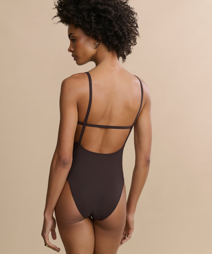 A woman with short curly hair models the Tre One Piece, a brown swimsuit featuring thin straps and a low back, as she stands against a plain beige background, turning her head slightly to the left.