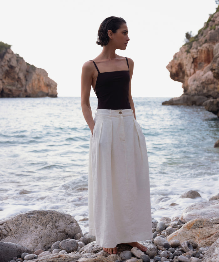A woman stands barefoot on a rocky beach, wearing the Tre One Piece with waves and cliffs behind her under a bright sky.