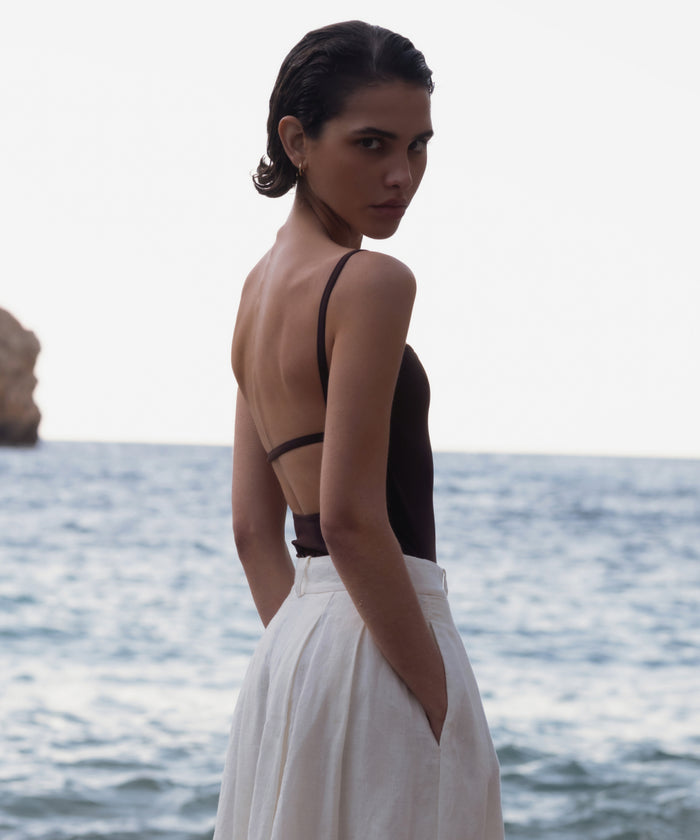 A person with short hair stands by the sea in a dark Tre One Piece, hands in pockets, glancing over their shoulder at the camera. The bright background shows sparkling water and a rocky formation.