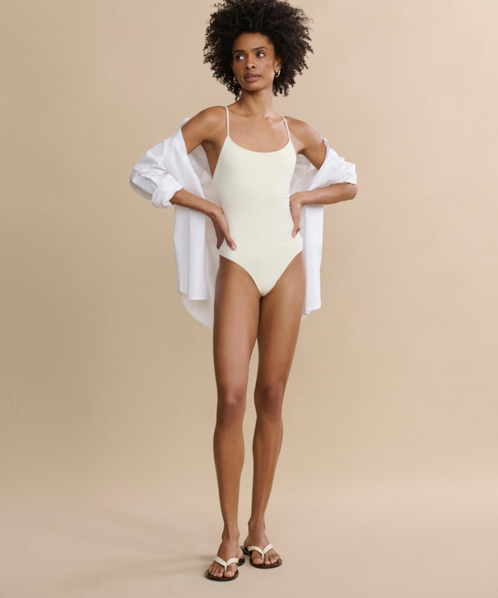 A woman with curly hair poses against a beige background wearing the Trentasei One Piece in light yellow, styled with a white unbuttoned shirt and white sandals, exuding classic style with one hand on her hip.