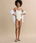 A woman with curly hair poses against a beige background wearing the Trentasei One Piece in light yellow, styled with a white unbuttoned shirt and white sandals, exuding classic style with one hand on her hip.