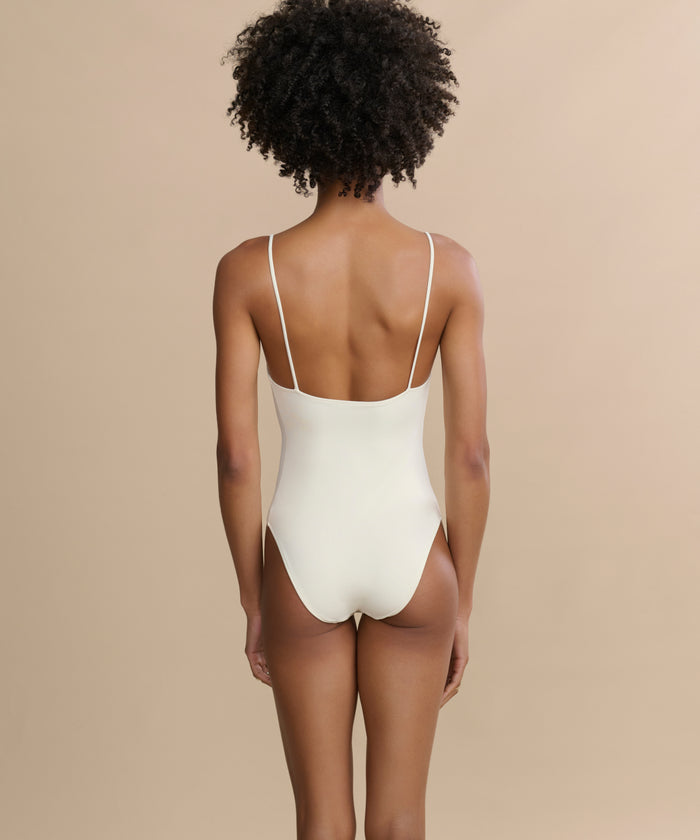 A person with short curly hair is seen from behind, wearing the Trentasei One Piece swimsuit with thin straps against a plain beige background.