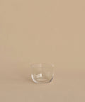 A clear, hand-blown tumbler from the Universal Low Glass Set of 4 rests on a beige surface with a matching background, highlighting Vincent Van Duysen’s minimalist style.