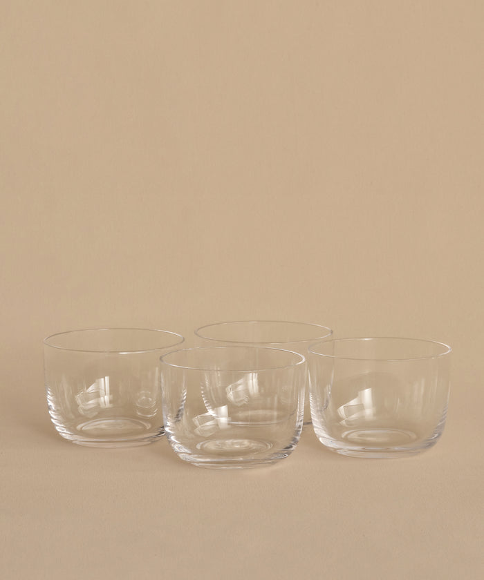 Four clear, round hand-blown bowls from the Universal Low Glass Set of 4 sit in two rows on a beige surface and background. The empty potassium glass bowls feature smooth, minimalist designs inspired by Vincent Van Duysen.